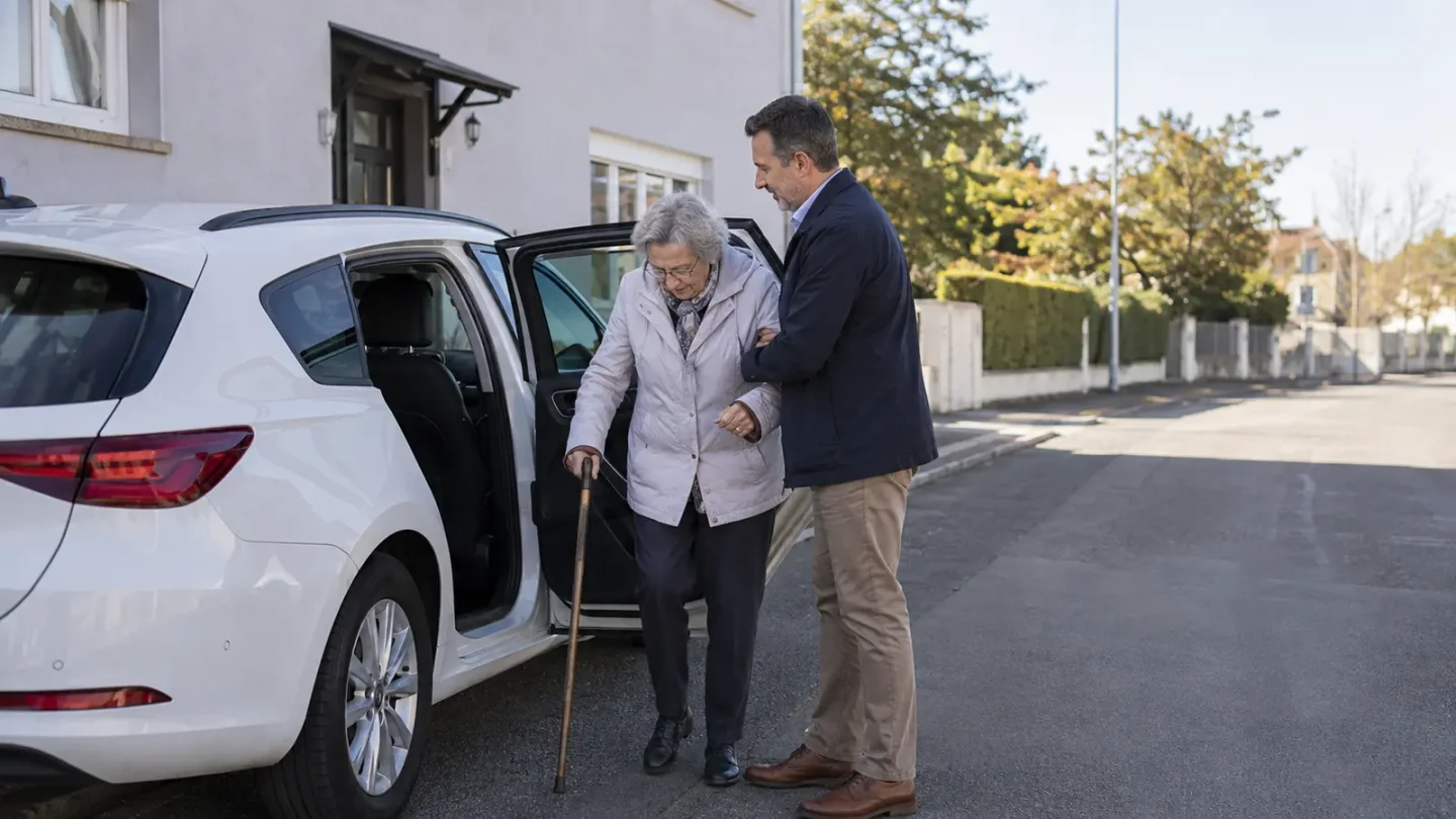 Une femme âgée est accompagnée par un conducteur à proximité d’un véhicule utilisé pour un transport médical.
